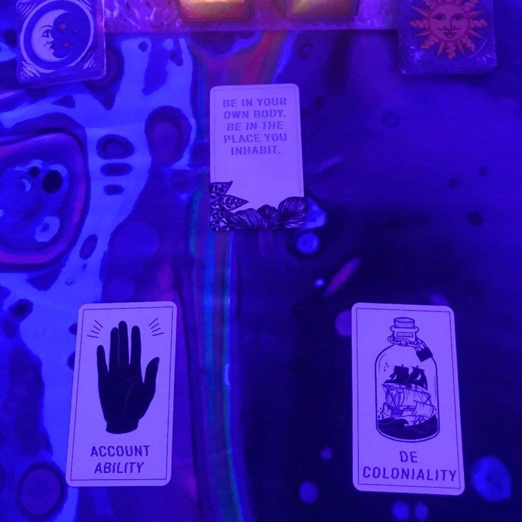 Three cards under blacklight,  At the top, a card with the words "Be in your own body.  Be in the place you inhabit." On the bottom left, a hand and the word Accountability.  On the bottom right, a ship in a bottle with a lock on it, and the word Decoloniality. 