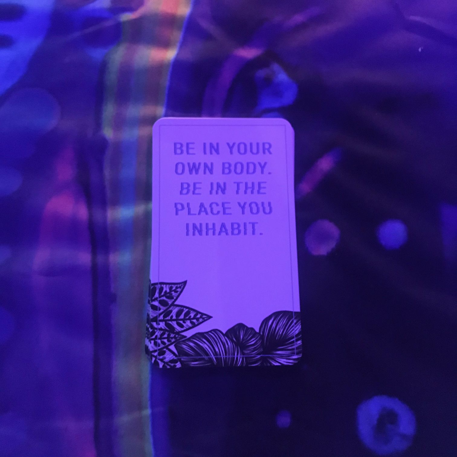 A card on a colorful reading cloth, under black light, with the words "Be in your own body.  Be in the place you inhabit."